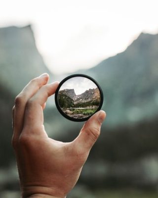 blurry background in nature with a hand holding up a lens that shows a clear picture demonstrating the power of awareness.