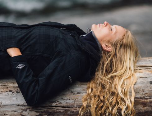 woman with long hair in a warm jacket laying on her back on a log with eyes closed to pause