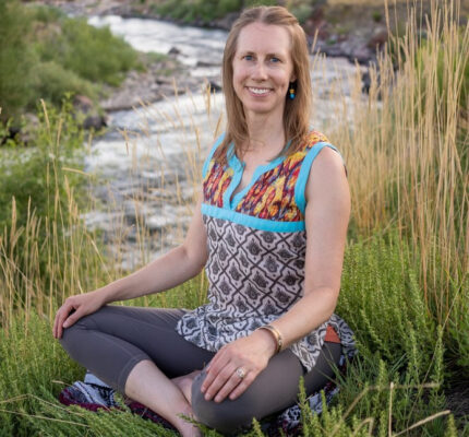 Author sitting cross-legged by a river in some tall grass and smiling at you