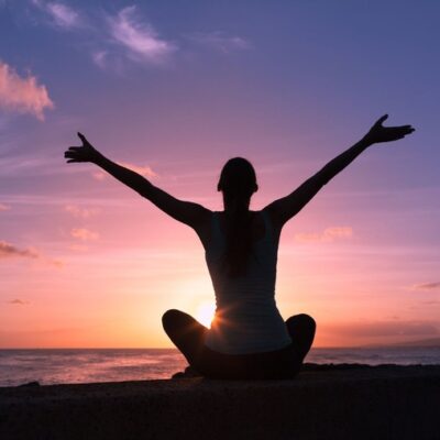 Woman sitting cross legged with arms outstretched towards an ocean sunset