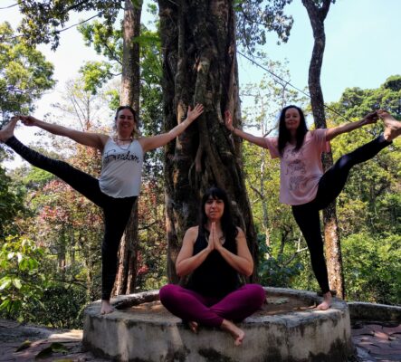 Three women in yoga poses in front of a tree. Two on the side in a balance pose, one sitting cross-legged between them.