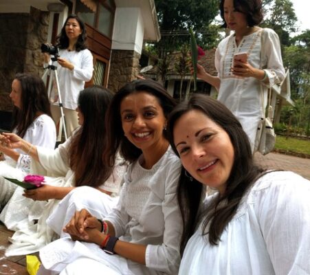 Two women smiling at the camera surrounded by other women sitting dressed in white for a ceremony