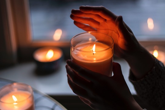 hands holding a glass candle with three other candles burning in the background with reflections on a window