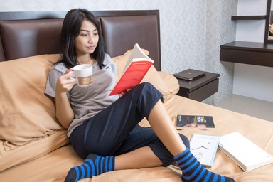 asian woman sitting on bed reading a book and drinking tea relaxing