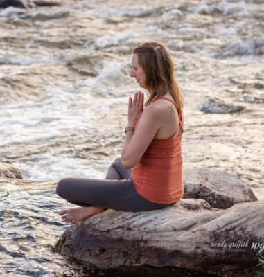 Author sitting cross-legged on a rock by a river with hands at her heart and eyes closed in silence with herself