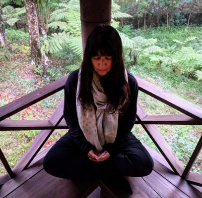 Woman sitting cross-legged, eyes closed in reflection on a platform surrounded by forest