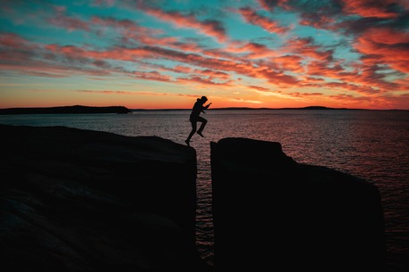 person jumping over a space between two rocks above the ocean at sunset