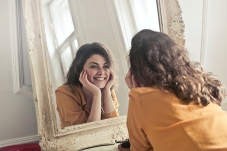 woman in gold shirt looking at herself in a mirror smiling with chin resting on the palms of her hands