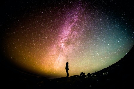 person standing looking up at a night sky with milky way enhanced with many colors representing the vastness of time