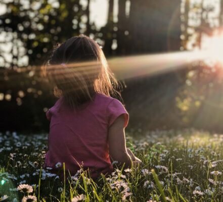 girl sitting and pausing in a field of daisies facing the sun with a sunbeam on her