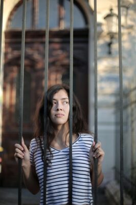 Woman peering out from a metal bar fence representing being prisoner despite being well-to-do