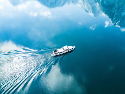 looking down on a moving white boat with the reflections of mountains in the water