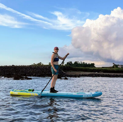 author on paddle board in ocean with rocks and green shore in background