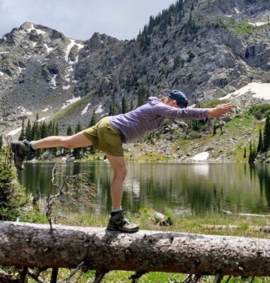 author on warrior three pose on a log with alpine lake and mountains in the background