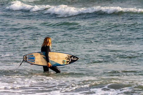 woman in a wetsuit with surf board walking out into the waves of the blue ocean