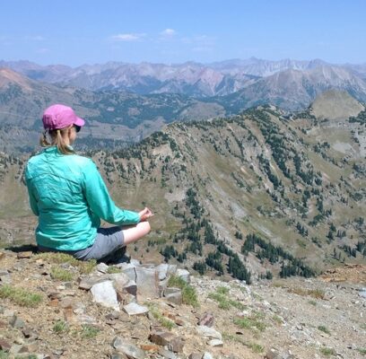 Woman practicing calm patience while sitting on top of a mountain overlooking a wide view into the valley helps to let go of outcomes